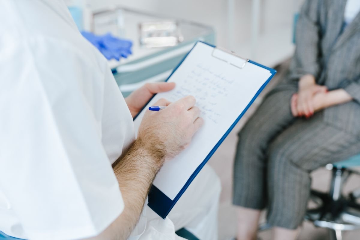 Clinician in a white coat writing notes on a blue clipboard while a seated patient in grey trousers waits nearby