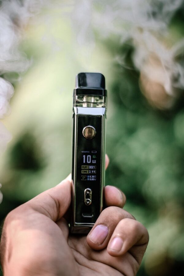 Hand holding a chrome pod-mod vape with a lit wattage display against a blurred outdoor background
